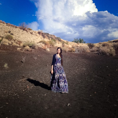 A woman wearing a long dress walks down a hill. Behind the hill are bright blue skies with lots of clouds.